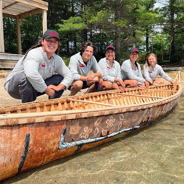 Five Indigenous youth squat beside a traditional birch-bark canoe.