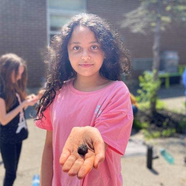 A young girl wearing a pink shirt stands by an urban outdoor garden holding a seed in her hand. 
