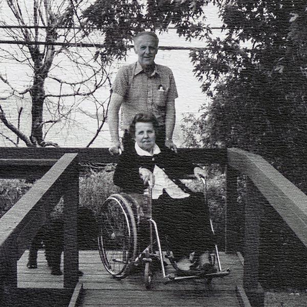 Image en noir et blanc d’une femme en fauteuil roulant en haut d’une rampe d’accès et d’un homme debout derrière elle.  