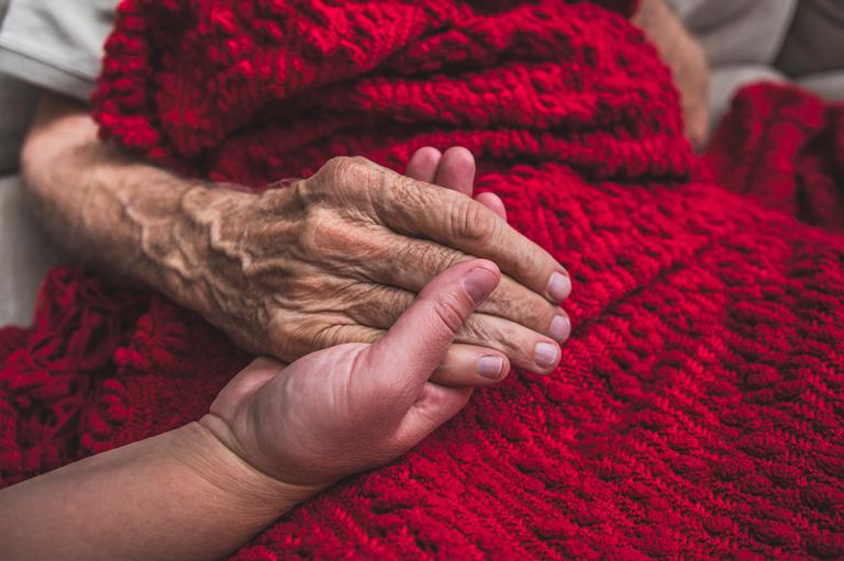 An elderly patient in hospice care holds a loved one’s hand.