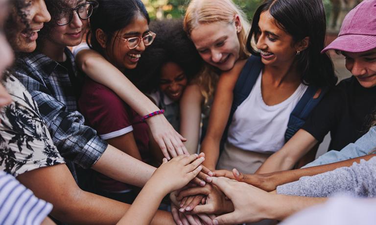 A group of teenagers smiling cheerfully outdoors while putting their hands together in a huddle. 