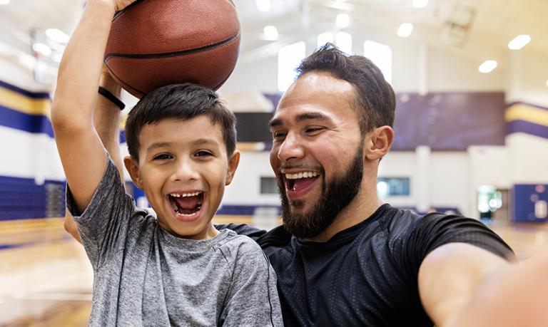 A father takes selfie while his son holds a basketball on his head.